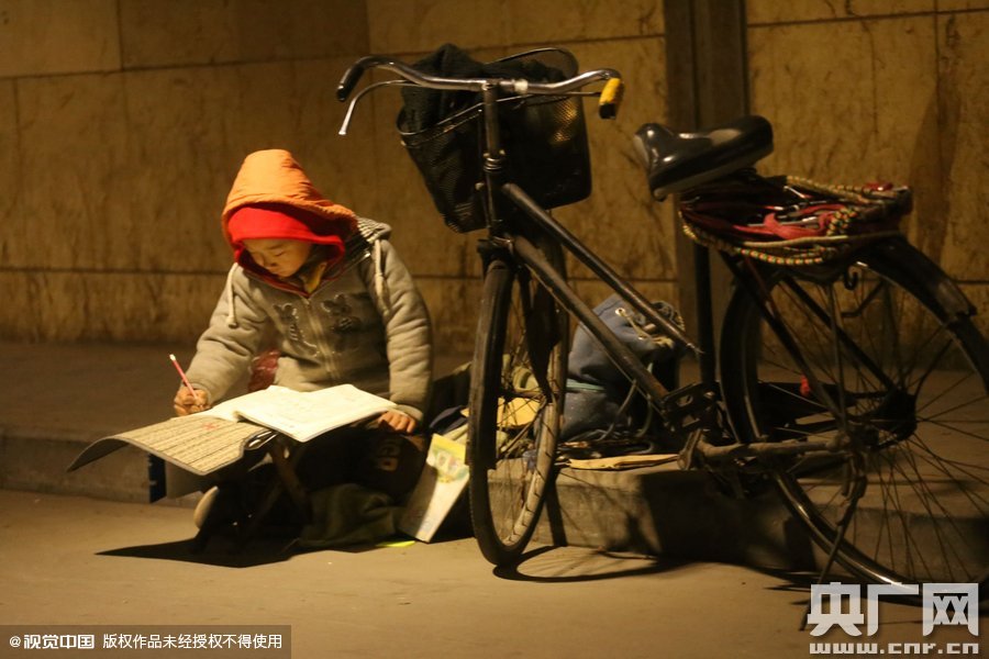 鄭州中原路京廣鐵路大橋下涵洞的馬路牙子邊，一位戴著紅色辣椒帽的小男孩趴在一個(gè)小馬扎上，上面鋪著一張車墊，在微弱的燈光下寫作業(yè)